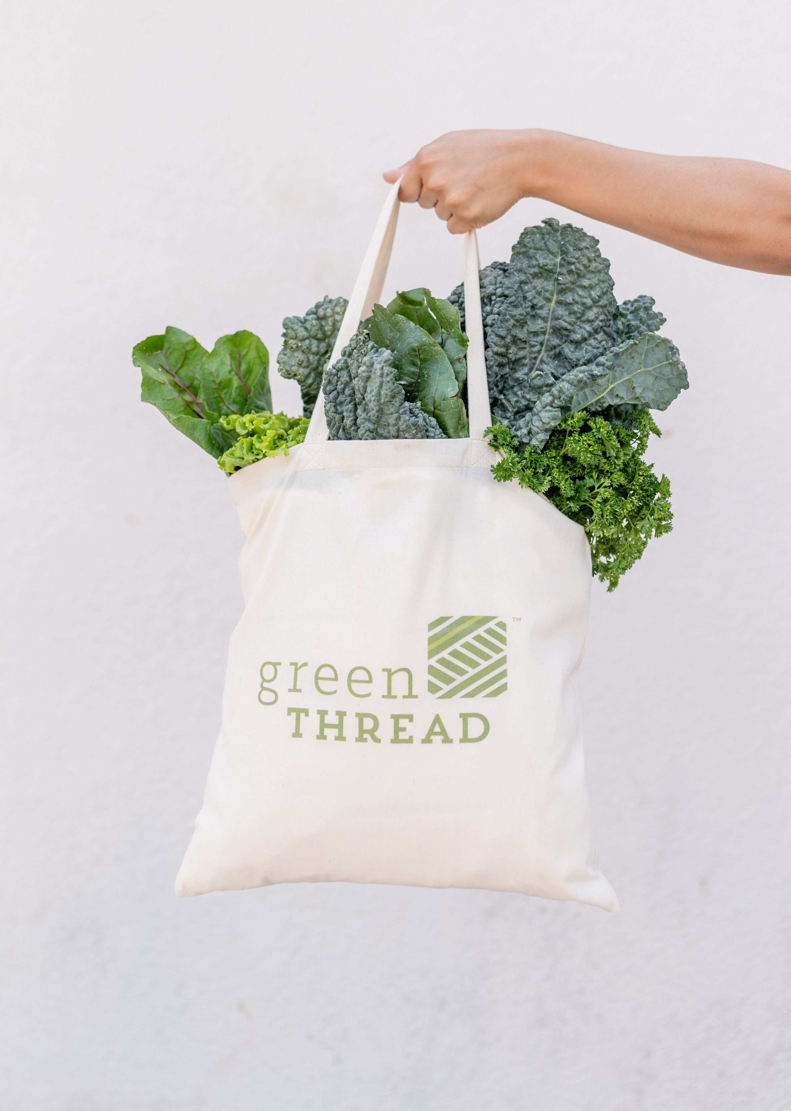 Image of hand holding tote bag filled with green vegetables with the Green Thread logo on it