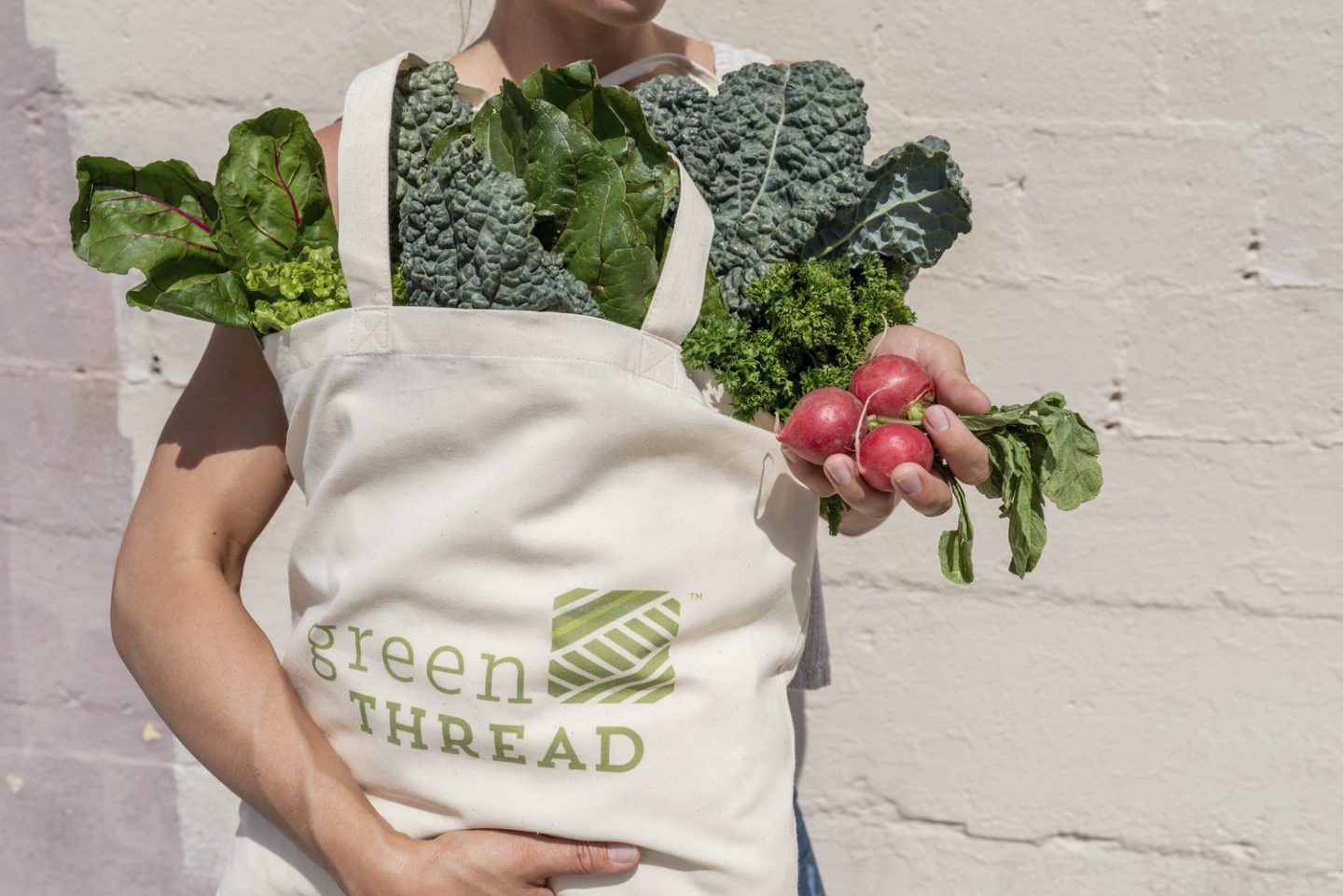 Image of person holding tote bag filled with green vegetables with the Green Thread logo on it. In the person's hand is a group of three radishes.
