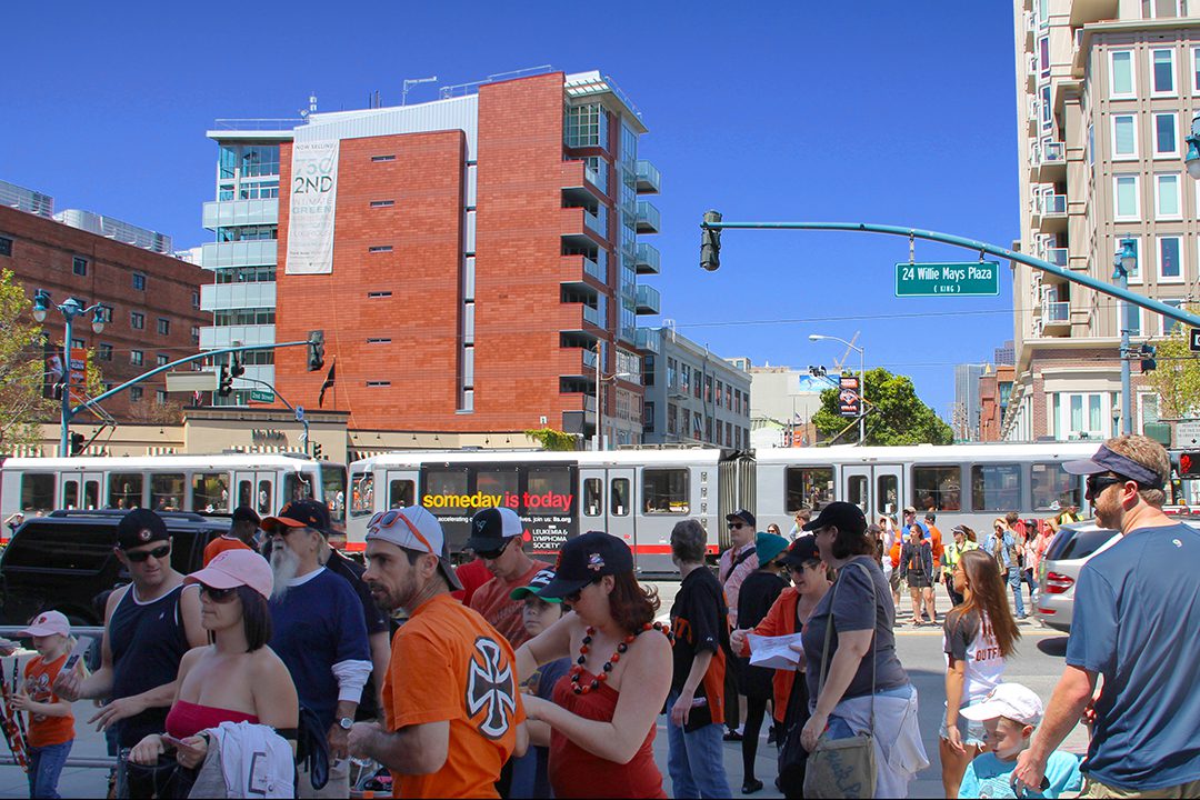 Image of a crowd of people outside of the San Francisco Giants' waterfront Oracle Park looking towards the 750 2nd Street residences while a train goes by the SFMTA Second Street & King Street light rail platform