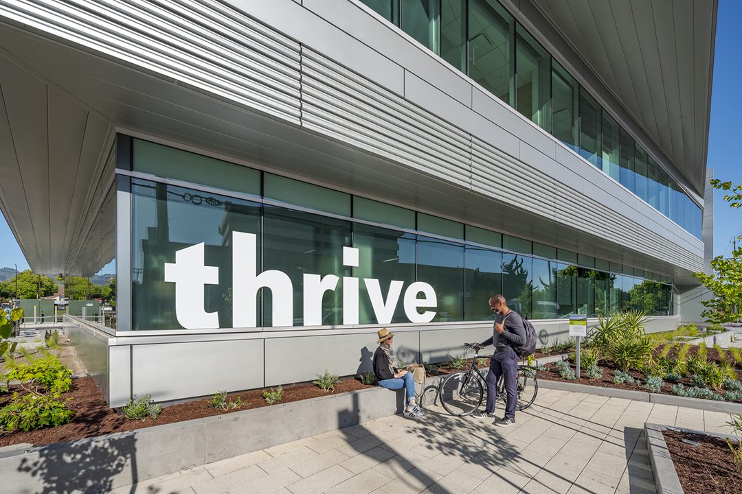 Image of the public pocket park on the corner of Parker & Tenth Street with woman sitting conversing with a man with bicycle standing in front of a window sign that says "thrive"