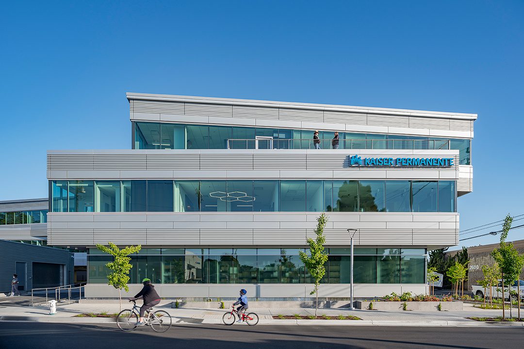 View of the Kaiser Permanente Medical Offices from Parker Street with bicyclists riding by and two people on the rooftop terrace
