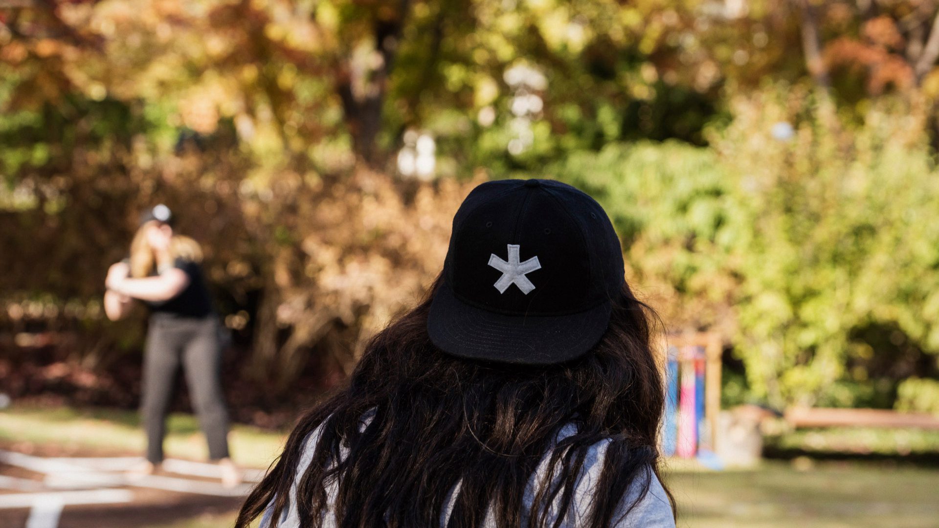 Image of a woman wearing a hat with the Multistudio asterisk on it in the foreground with another woman at-bat in softball in the background. The background is blurred a bit but one call tell it is a beautiful, sunny, autumn day.