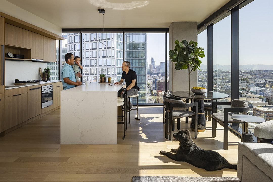 Image of a man standing holding a baby while another man sits across from him at the kitchen island. A black dog lays in the sun on the floor.