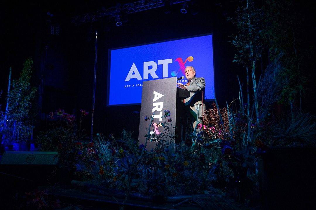 An event speaker behind a podium with the ARTx logo on the front and on projection screen behind.